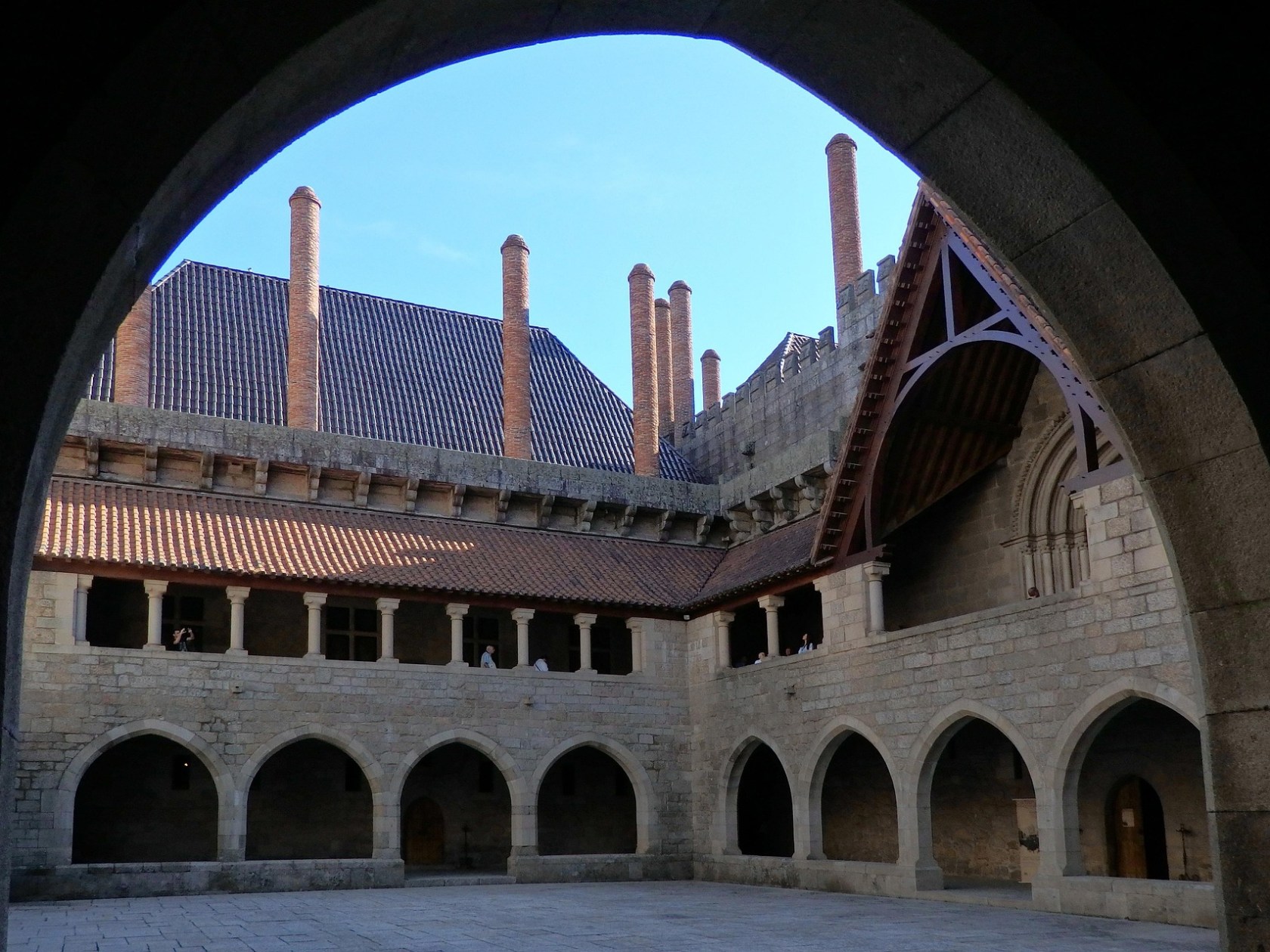 Stone courtyard with arched walkways and tall brick chimneys against a blue sky.