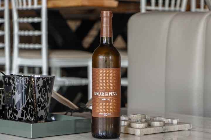 Wine bottle on table with glass and chairs in background.