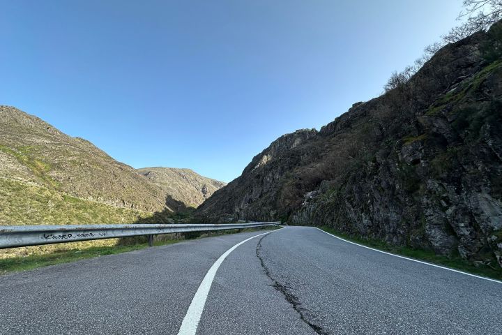 Curved road with guardrail beside rocky hills and clear blue sky.
