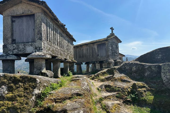 Stone granaries on stilts with a cross on a rocky hill under a clear blue sky.
