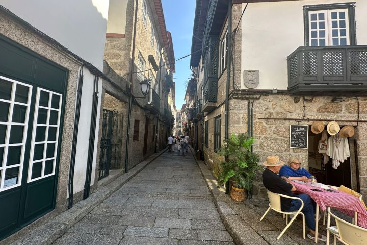 Narrow European street with people sitting at a café, cobblestone path, and stone buildings.
