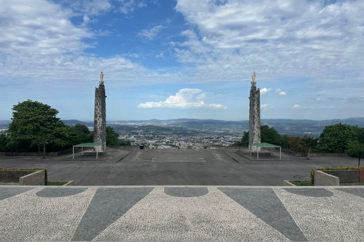 Wide view of open plaza with two tall statues, cityscape and mountains in the background under a cloudy sky.