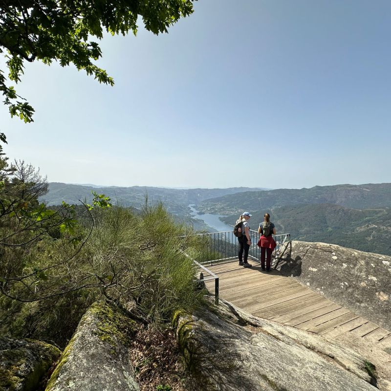 Three people on a wooden platform overlooking a mountainous landscape with a river.